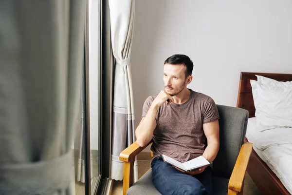 Pensive young man sitting in armchair in the morning, looking through window and filling gratitude journal