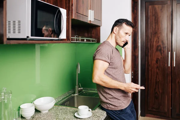 Vážný Mladý Muž Opírající Kuchyňský Pult Když Mluví Telefonu Příbuzným — Stock fotografie