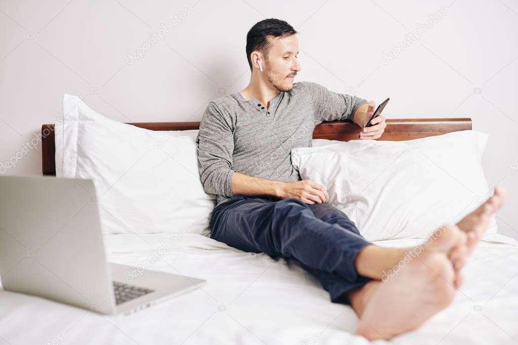 Barefoot young man resting on bed and reading article on screen of his smartphone