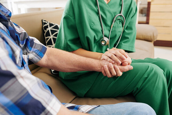 Крупный план медсестры в зеленой форме, держащейся за руку пожилого пациента при посещении его дома и рассказывающей о процессе выздоровления
