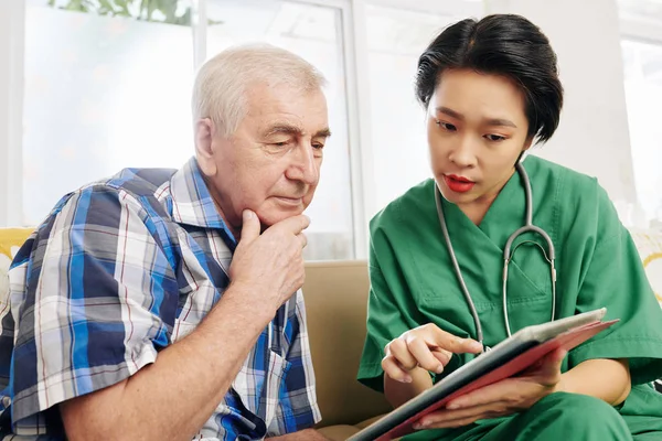 Concerned senior man reading medical tests results on talbet computer in hands of nurse visiting him