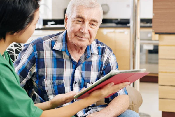 Smiling senior man reading information on tablet computer in hands of nurse visiting him at home