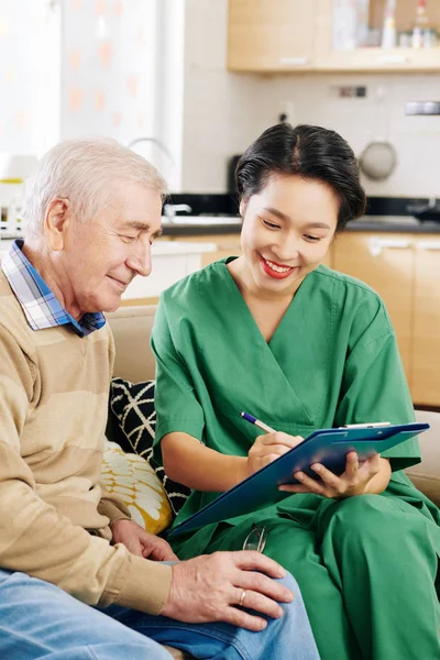 Young Smiling Medical Nurse Writing Complaints Senior Man Visiting Him — Stock Photo, Image