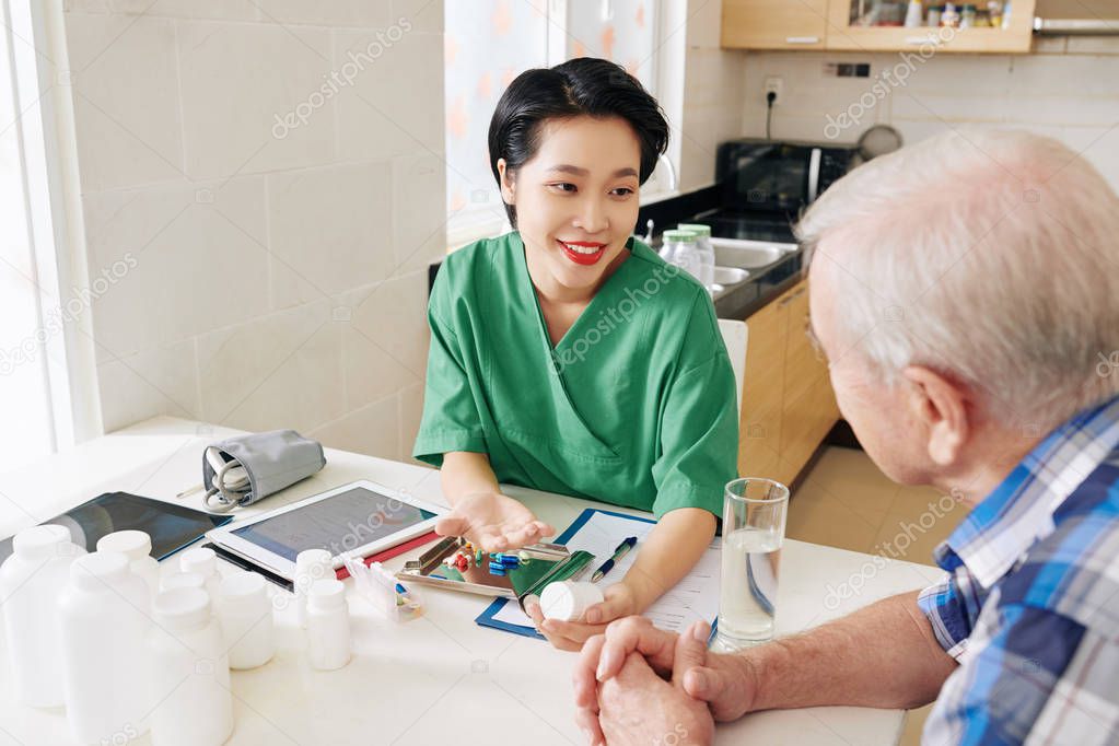 Smiling Vietnamese medical nurse visiting senior patient to check his health status and explain what medicine he should take daily