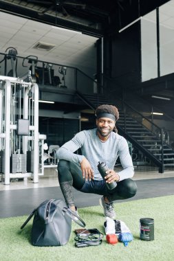 Spor beslenme çantası ve spor aksesuarlarıyla spor salonunda oturan neşeli fitness antrenörü.