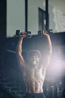 Gömleksiz, mutlu, zinde bir adamın portresi kafasının üzerine ağır halterler kaldırıyor.