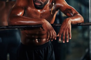 Spor salonundaki yoğun eğitimden sonra trabzanlara yaslanan terli kaslı adamın kırpılmış görüntüsü.