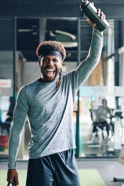 Excited happy young man rising hand with bottle of fresh water and shouting after intense training in gym