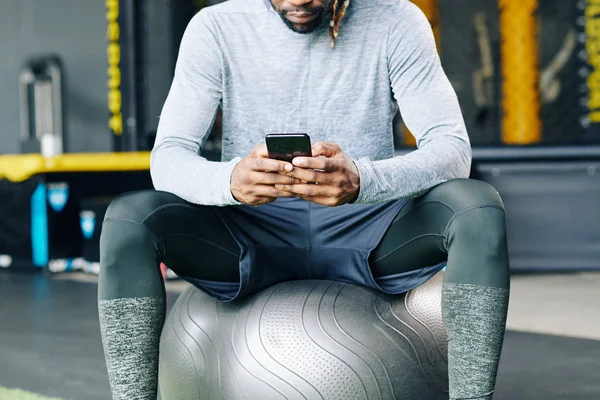大きなフィットネスボールに座っている若いスポーツマンの作物のイメージとスマートフォン上で健康アプリケーションをチェック — ストック写真