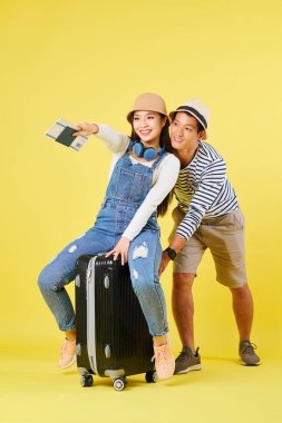 Genç Vietnamlı bir adam bavulu iterken kız arkadaşı üzerine oturur ve elinde pasaportlarla uçak biletleriyle işaret eder.