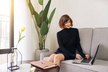 Siyah elbiseli güzel bir bayan kanepede oturuyor ve bilgisayarındaki e-postaları cevaplıyor.