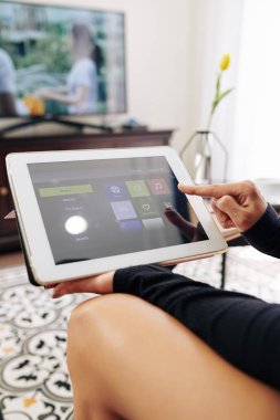 Genç bir kadının akıllı televizyondaki film, şov veya müziği seçmek için tablet bilgisayarında medya hizmeti uygulamasını kullanışının yakın plan görüntüsü