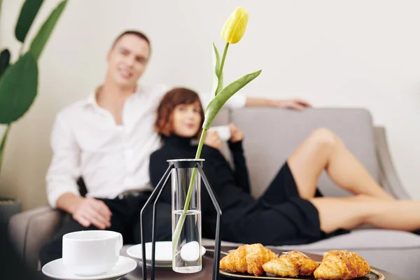 在沙发上休息的年轻夫妇客厅里 桌上放着郁金香花 咖啡杯和羊角面包 — 图库照片