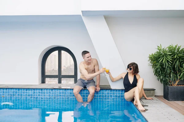 快乐的年轻夫妇坐在游泳池边 边喝着果子酒边碰杯 — 图库照片