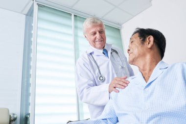 Gülümseyen pratisyen doktor iyileşme terapisi sırasında ona destek olmak için Vietnamlı bir adamın omzunu okşuyor.