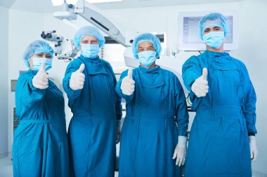 Team of surgeons in scrubs and face masks showing thumbs-up after suvcccessful surgery clipart
