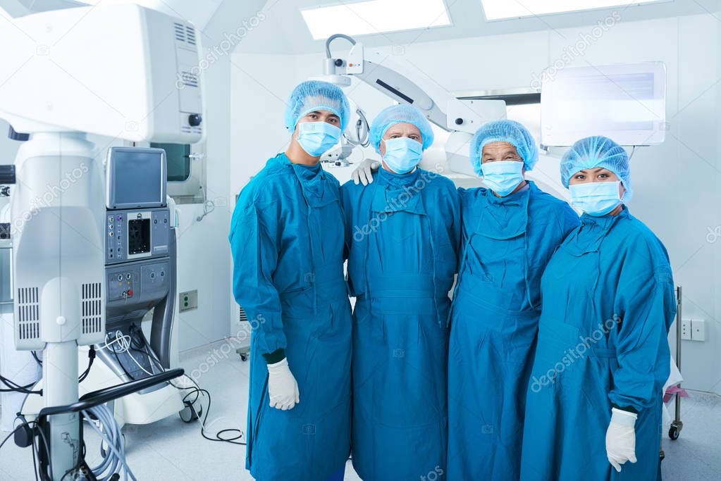 Hugging surgical team in scrubs standing in operating theater after surgery and looking at camera