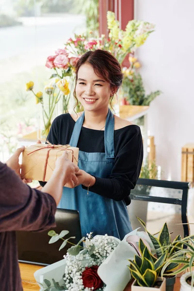 Cheerful pretty Vietnamese gift shop worker giving wrapped present to customer
