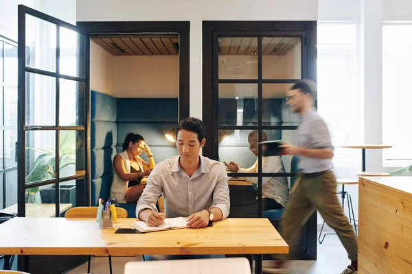 积极的年轻亚洲企业家坐在办公室桌旁 当他的同事们在幕后工作时 他们会在计划书中写下自己的想法 — 图库照片