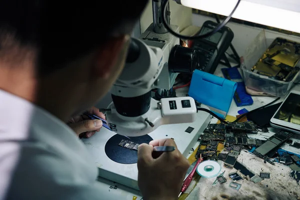 修理工坐在桌子旁 手里拿着坏了的智能手机零件 用显微镜观察着逻辑板 — 图库照片