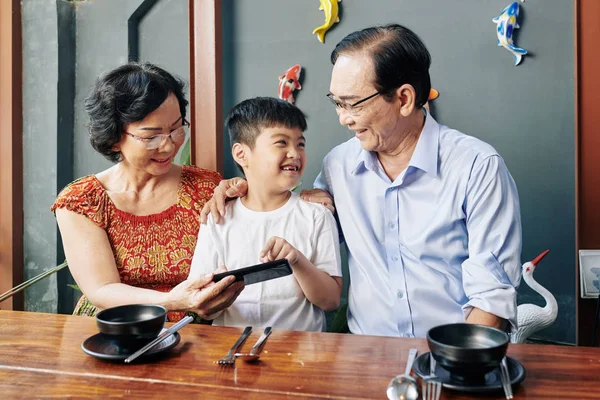 Happy Vietnamese Boy Showing Game Smartphone Screen Grandparents Sitting Cafe — Stok fotoğraf