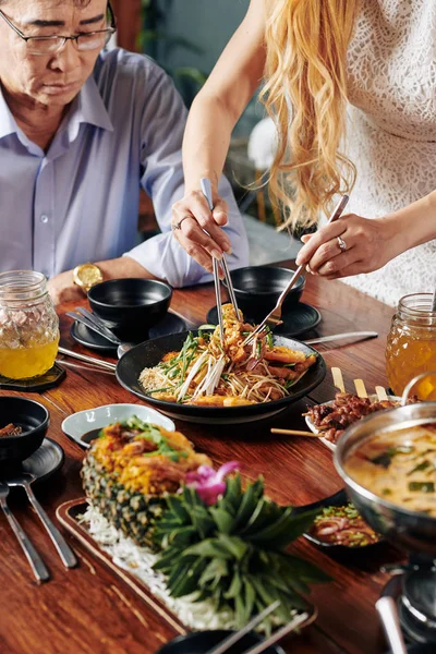 Housewife using fork and pliers when putting delicious Vietnamese dish from bowl in her plate and plate of her father