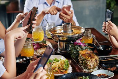 Hands of party guests sitting at dinner table with traditional Asian cuisine and texting friends of checking social media on smartphones