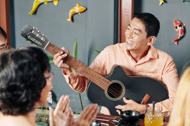 Happy middle-aged Asian man playing guitar and singing a song for his relatives at family dinner