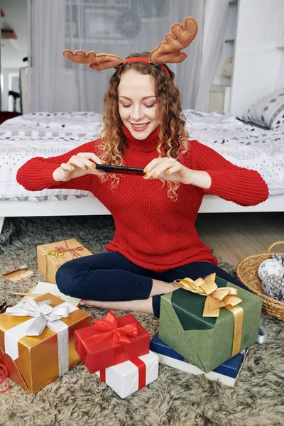 彼女はクリスマスのお祝いのために包まれたプレゼントの写真を撮るトナカイアントラーで若い白人女性を笑顔 — ストック写真