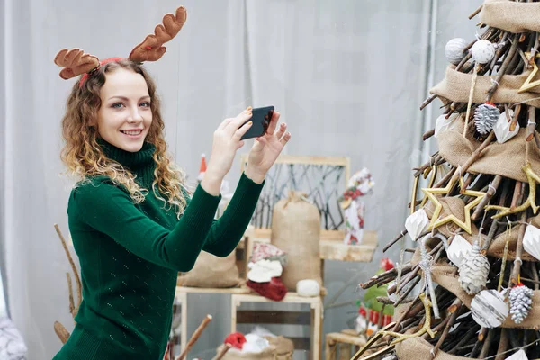 美しい若い笑顔の女性が木の枝で作られた装飾クリスマスツリーの写真を撮る — ストック写真