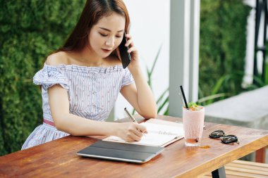 Yazlık elbiseli güzel Vietnamlı bir kadın kafeterya masasında not alırken telefonla konuşuyor.