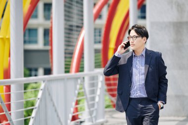 Gözlüklü ciddi bir Asyalı girişimci dışarıda yürüyor ve iş ortağıyla telefonda konuşuyor.