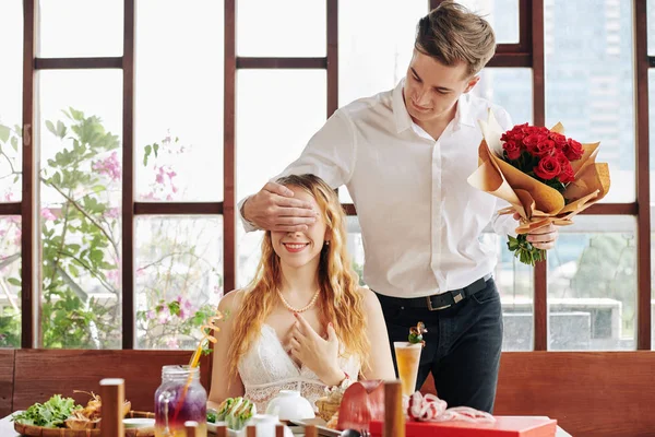 Smiling attractive young man with bouquet of fresh roses in hand covering eyes of his happy girlfriend sitting at restaurant table