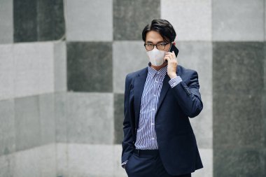 Yüz maskesi takmış ciddi bir Asyalı girişimci telefonla konuşuyor.