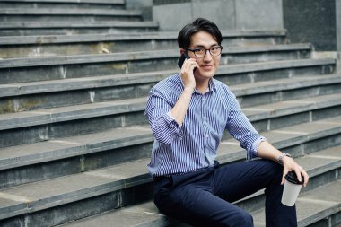 Pozitif Asyalı genç bir adamın portresi açık havada tek kullanımlık kahveyle oturuyor ve telefonda konuşuyor.