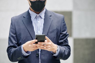 Koruyucu maskeli iş adamının iş arkadaşıyla mesajlaştığı ya da sosyal medyayı kontrol ettiği kırpılmış görüntü