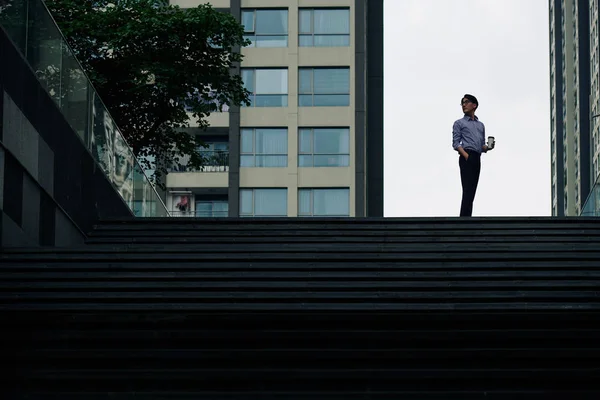 階段の上に立ってコーヒーを飲んでオフィスの建物を見るために背を向ける男 — ストック写真