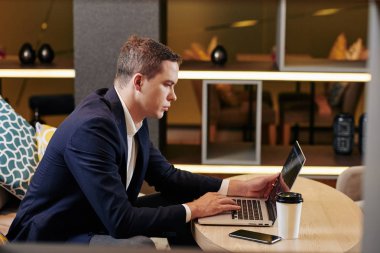 Ciddi genç ve yakışıklı girişimci kafeterya masasında bir fincan kahveyle oturuyor ve dizüstü bilgisayarla çalışıyor.