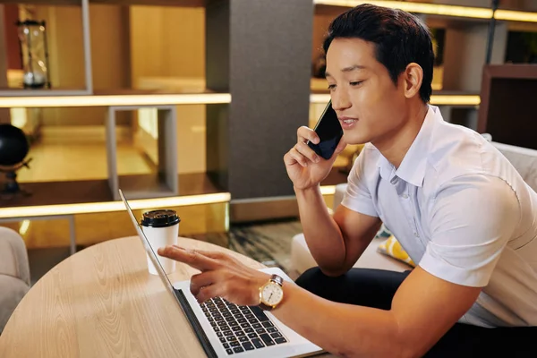 Young Vietnamese businessman pointing at laptop screen and specifying project details when talking on phone with client