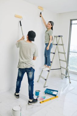Kot pantolon ve tişört giyen neşeli Vietnamlı çift odaların duvarlarını beyaz boyayla boyuyor.