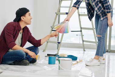 Gülümseyen Asyalı genç adam kız arkadaşının elindeki kumaş parçalarıyla rengini gösteriyor ve yatak odasının duvarları için gölgeyi seçiyor.