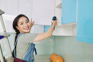 Mutfağındaki dolabın kapısını tamir ederken elektrikli tornavida kullanan neşeli, güzel Vietnamlı genç kadın.