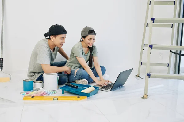 ツールや塗料で新しいアパートの床に座ってキャップで陽気な若いアジアのカップルやデザインのアイデアをオンラインで検索 — ストック写真