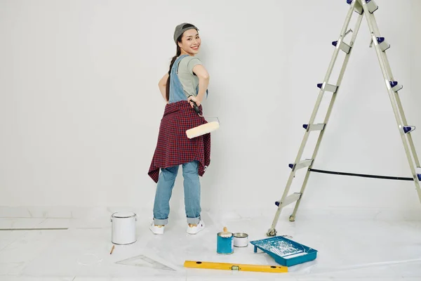 Cheeful young Vietnamese woman with roller in hand turning back and looking at camera after painting wall