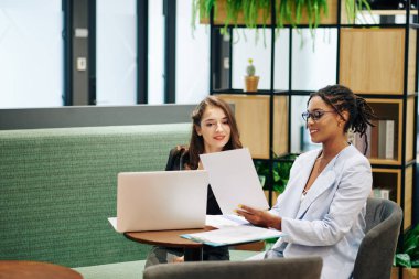 Gözlüklü, gülümseyen iş kadını toplantıda genç meslektaşına mali raporla birlikte belge gösteriyor.