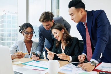 Multi-ethnic business people smiling when looking at screen of smartphone in hand of young coworker