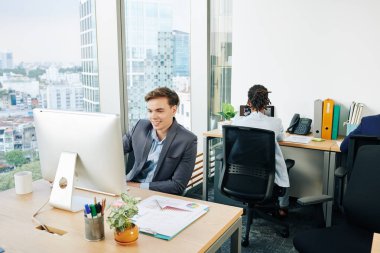 Smiling handsome young successful businessman sitting at office table and reading e-mails on computer screen