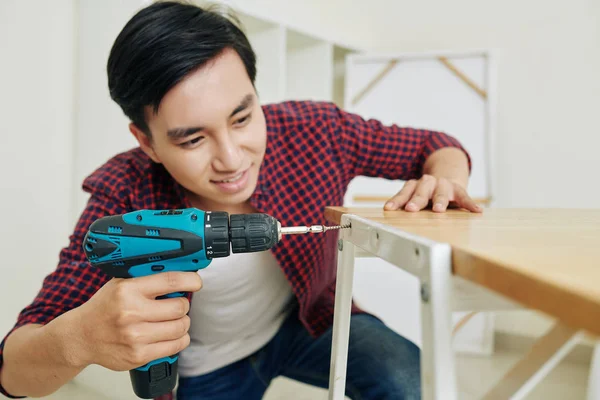 正の若いベトナム人男性が新しいアパートで家具を組み立て 電動ドライバーでネジを締めます — ストック写真
