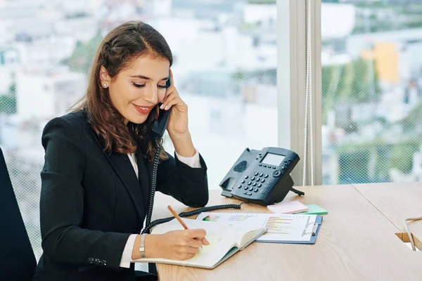 Smiling Pretty Young Office Administrator Talking Phone Writing Details Notepad — Stockfoto
