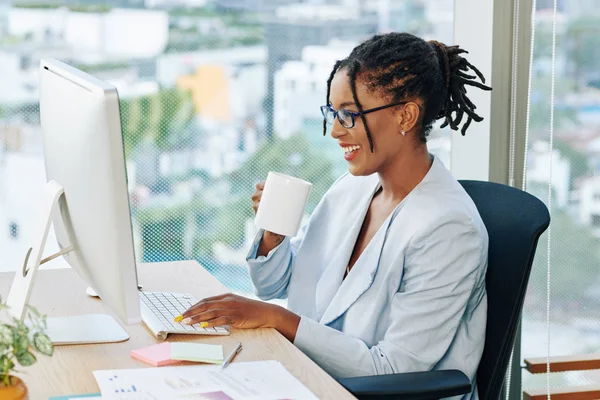笑着漂亮的女商人 可怕地喝着咖啡 在电脑屏幕上读着同事们的有趣信息 — 图库照片